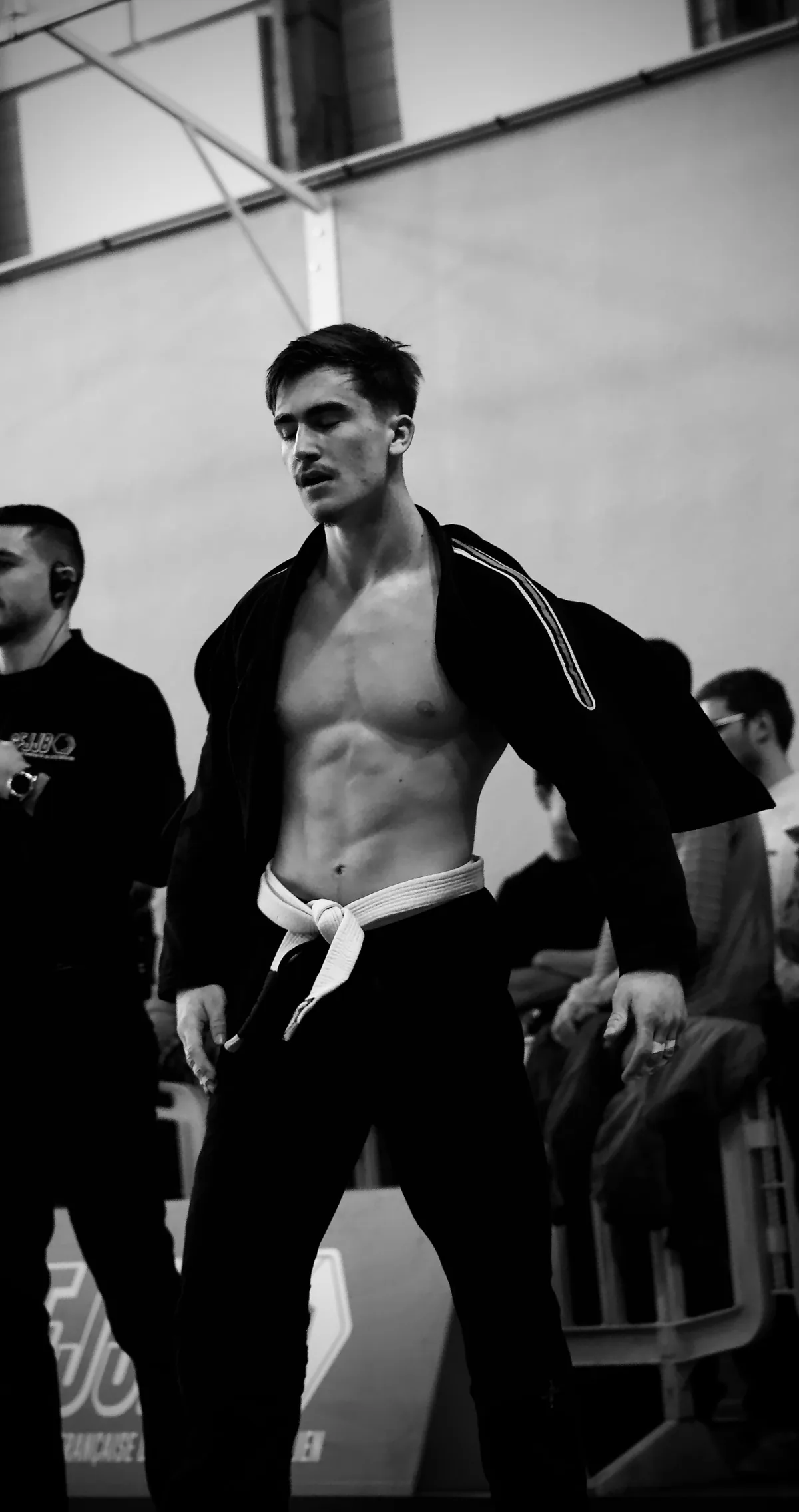 Photographie en noir et blanc d’un compétiteur de jiu-jitsu brésilien, kimono ouvert et ceinture blanche, debout dans une salle de compétition.