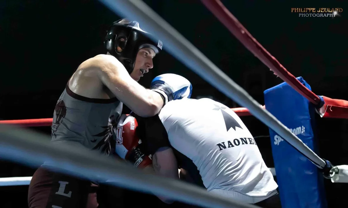 Deux boxeurs casqués au corps à corps dans les cordes d’un ring, pendant une reprise.